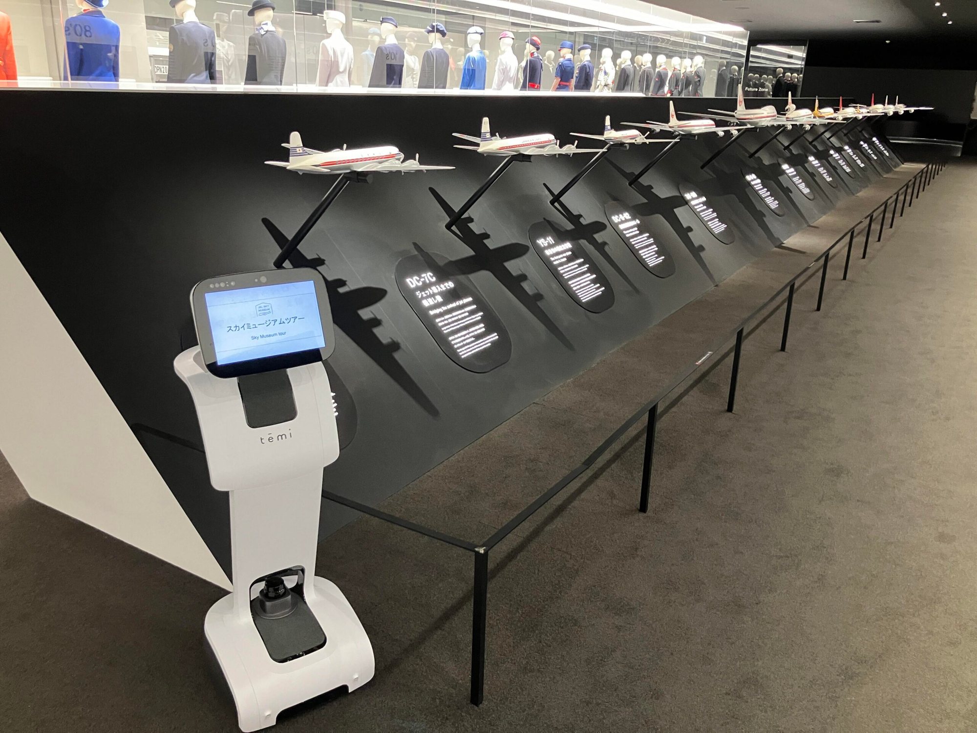 JALスカイミュージアムの展示室。歴代機の模型が並ぶ黒い壁の手前に、ツアーの案内画面を出した白い temi が立っている