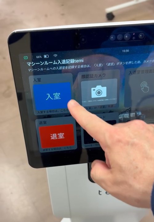 「入室」「退室」「顔認証カメラ」のボタンが並ぶ temi の画面に、指が触れている