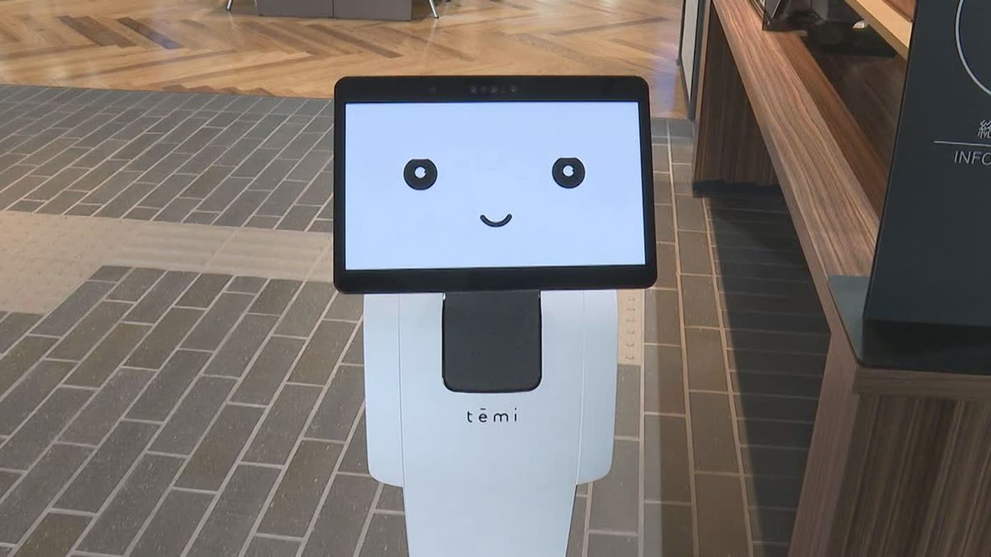 笑顔を表示した白い temi の上半身。奥に木のカウンターと INFORMATION の案内板がある