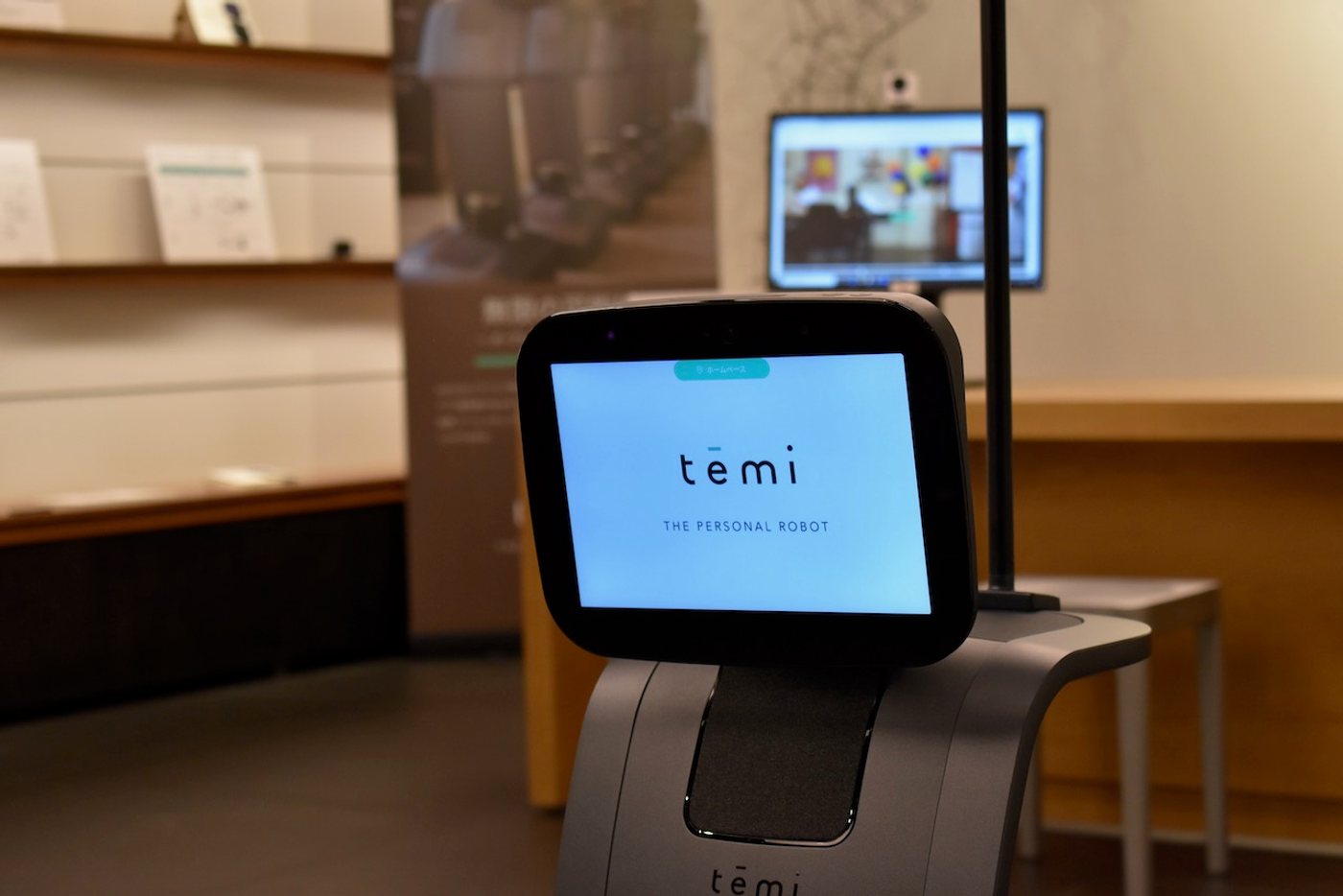 実機の temi。上部の画面に temi のロゴと「THE PERSONAL ROBOT」の文字が表示され、後方の棚には製品の解説パネル、右奥にはモニターが置かれている